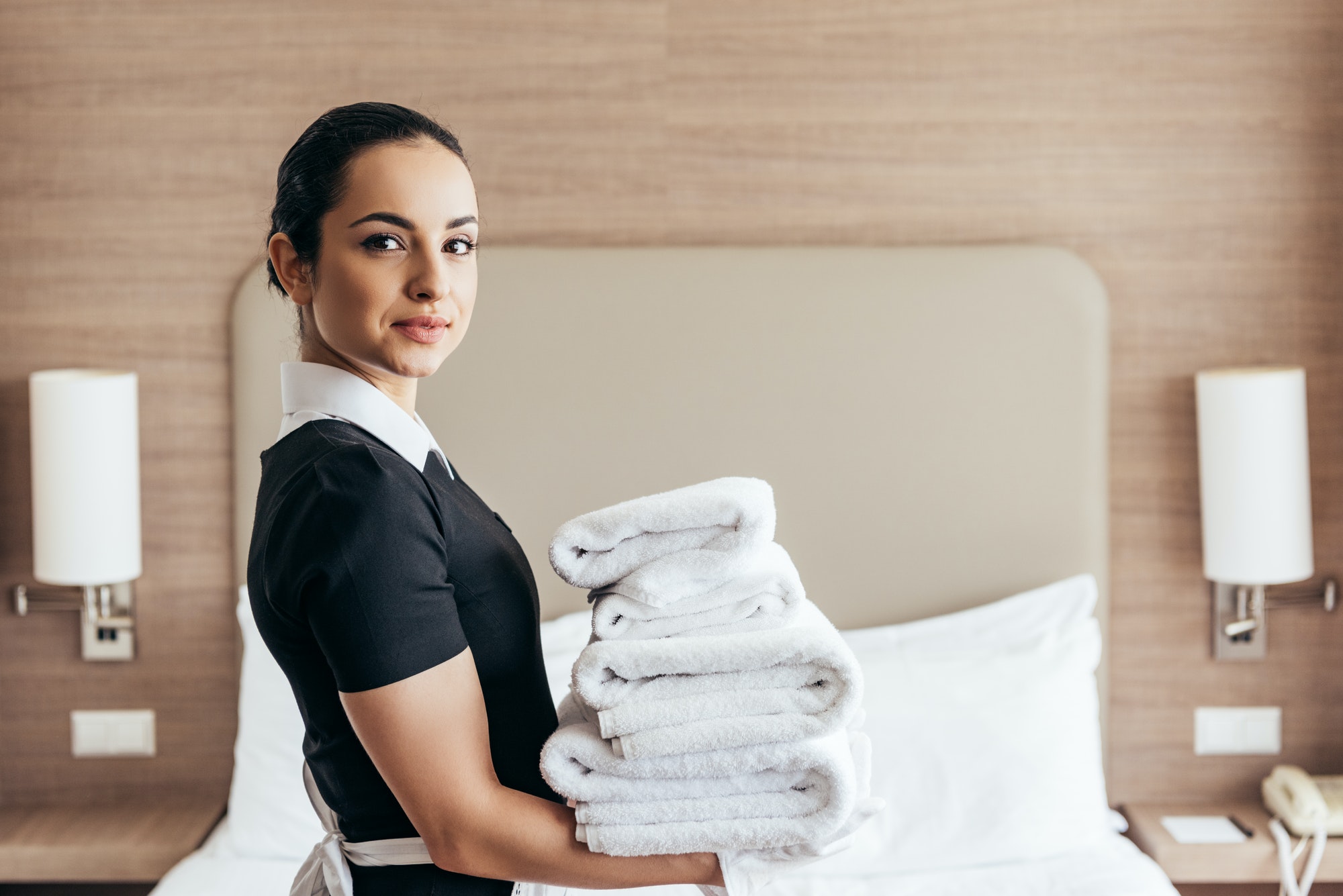 smiling maid holding pile of folded towels near bed and looking at camera in hotel room.jpg